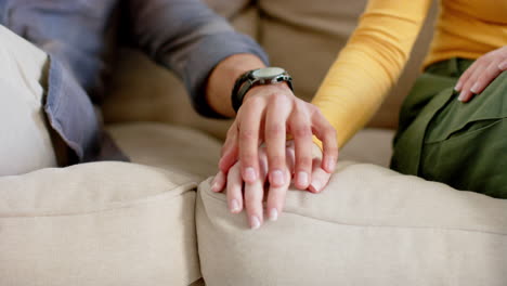 Midsection-of-diverse-couple-sitting-on-sofa-holding-hands-at-home,-in-slow-motion