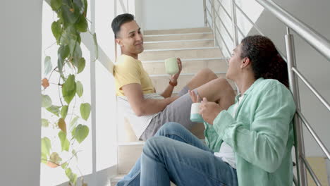 Happy-diverse-gay-male-couple-sitting-on-sunny-staircase-having-coffee-and-talking,-slow-motion