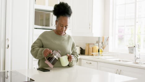 Glückliche-Afroamerikanische-Seniorin-Gießt-Kaffee-Ein-Und-Lächelt-In-Sonniger-Küche,-Zeitlupe