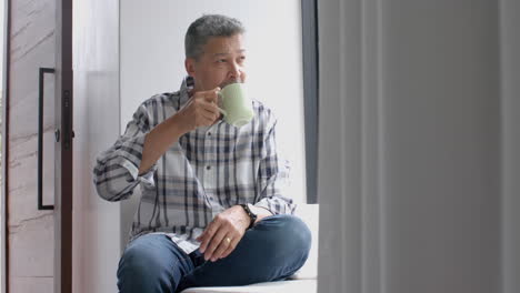 Happy-senior-biracial-man-sitting-at-window-and-drinking-coffee-at-home,-copy-space,-slow-motion