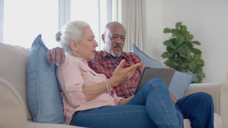 Happy-senior-biracial-couple-sitting-on-couch-and-using-tablet-at-home,-slow-motion