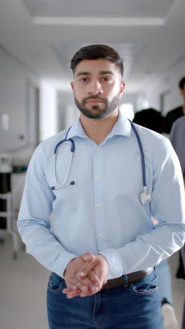 Vertical-video-of-portrait-of-happy-biracial-male-doctor-in-hospital-corridor,-slow-motion