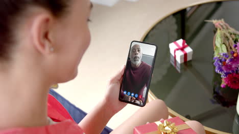 Biracial-woman-holding-smartphone-with-african-american-man-with-gift-on-screen-with-gift-on-desk