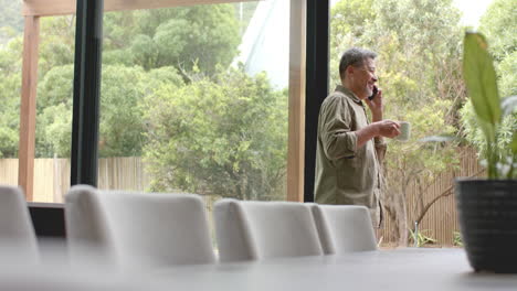 Happy-senior-biracial-man-by-window-talking-on-smartphone-holding-coffee,-copy-space,-slow-motion
