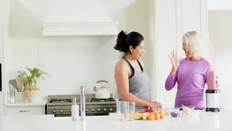 Two-happy-diverse-senior-women-preparing-cocktail-and-giving-high-five-in-kitchen,-slow-motion