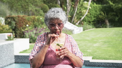 Happy-senior-african-american-woman-in-sun-by-pool-drinking-cocktail,-copy-space,-slow-motion