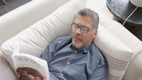 Focused-senior-biracial-man-in-glasses-lying-on-couch-reading-book-at-home,-slow-motion
