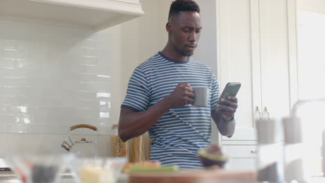 Feliz-Hombre-Afroamericano-Tomando-Café-Y-Usando-Un-Teléfono-Inteligente-En-Una-Cocina-Soleada,-Cámara-Lenta