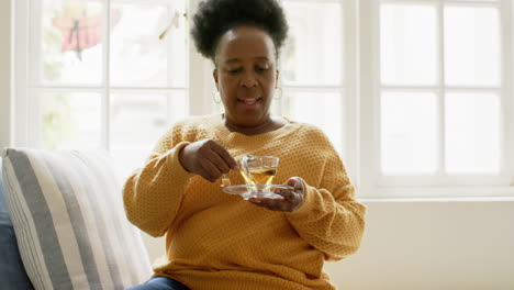 Glückliche-Afroamerikanische-Seniorin,-Die-Mit-Tee-Sitzt-Und-In-Einem-Sonnigen-Zimmer-Lächelt,-Zeitlupe