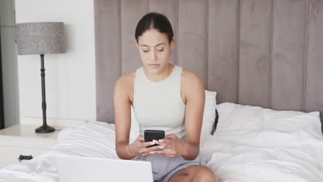 Biracial-woman-sitting-on-bed-using-laptop-and-smartphone,-slow-motion