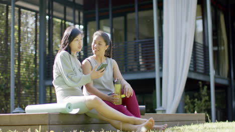 Glückliche-Asiatische-Freundinnen-Mit-Wasserflasche-Und-Smartphone-Lächeln-Auf-Der-Terrasse,-Zeitlupe