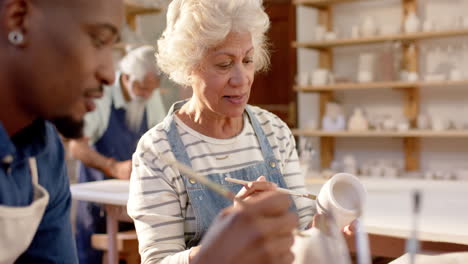 Two-diverse-male-and-female-potters-glazing-clay-jug-and-discussing-in-pottery-studio,-slow-motion