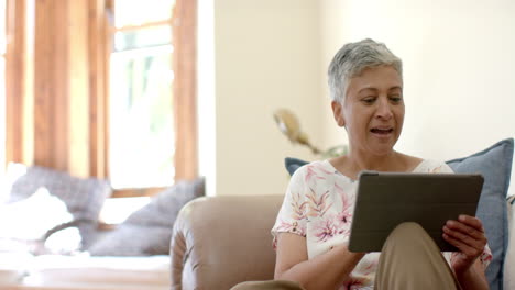 Happy-senior-biracial-woman-sitting-on-couch-laughing-and-using-tablet-at-home,-slow-motion