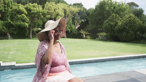 Glückliche-ältere-Afroamerikanische-Frau-Mit-Sonnenhut,-Die-In-Der-Sonne-Am-Pool-Sitzt,-Kopierraum,-Zeitlupe