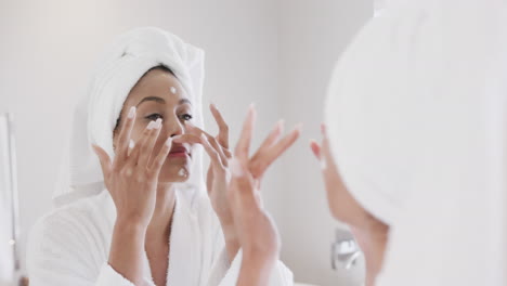 Biracial-woman-applying-face-cream-looking-in-mirror-in-bathroom,-slow-motion