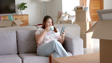 Plus-size-biracial-woman-on-sofa-using-smartphone-having-coffee-in-new-home,-copy-space,-slow-motion