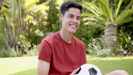 Happy-biracial-man-sitting-with-football-after-training-in-sunny-garden,-copy-space,-slow-motion