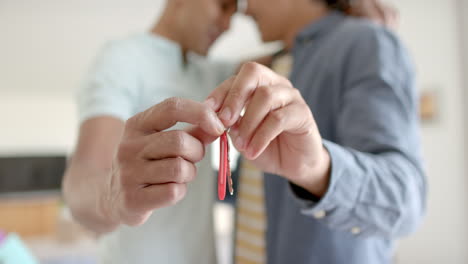 Hands-of-diverse-gay-male-couple-holding-keys-to-new-house,-embracing-in-living-room,-slow-motion