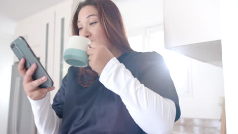 Happy-plus-size-biracial-female-doctor-having-coffee-using-smartphone-at-home,-slow-motion