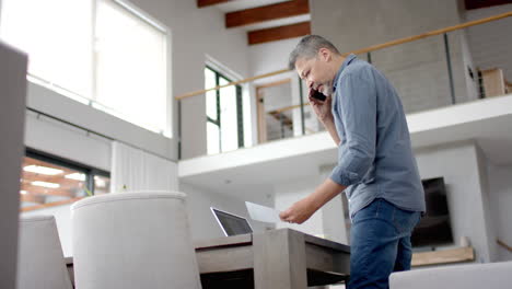 Senior-biracial-man-on-phone,-standing-looking-at-paperwork,-home-working,-copy-space,-slow-motion