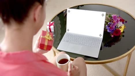 Caucasian-woman-holding-gift-and-using-laptop-with-copy-space-on-blank-screen