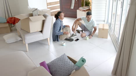 Happy-diverse-gay-male-couple-moving-into-new-home-sitting-on-floor,-copy-space,-slow-motion