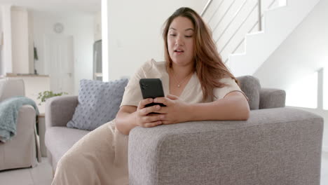 Happy-plus-size-biracial-woman-having-smartphone-video-call-sitting-on-sofa,-copy-space,-slow-motion