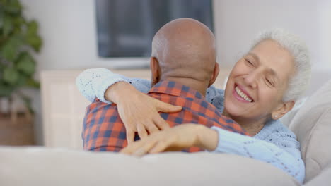 Happy-senior-biracial-couple-sitting-on-couch-and-embracing-at-home,-slow-motion