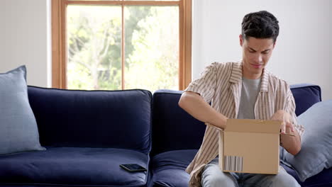 Happy-biracial-man-opening-delivery-in-cardboard-box-and-using-smartphone,-copy-space,-slow-motion