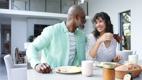 Happy-biracial-couple-having-scrambled-eggs-for-breakfast-and-talking-in-kitchen,-slow-motion