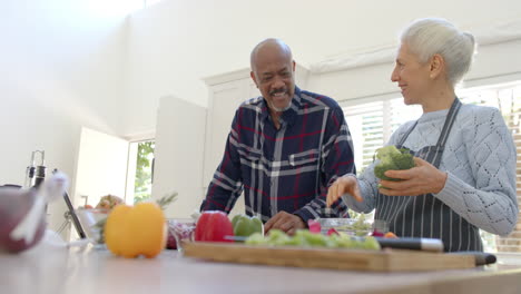 Happy-diverse-senior-couple-preparing-vegetables,-using-tablet-in-kitchen,-copy-space,-slow-motion