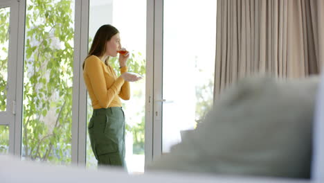 Thoughtful-caucasian-woman-drinking-tea-and-looking-through-window-at-home,-copy-space,-slow-motion