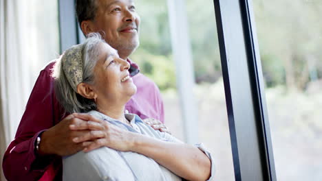 Happy-biracial-senior-couple-embracing-at-window-at-home