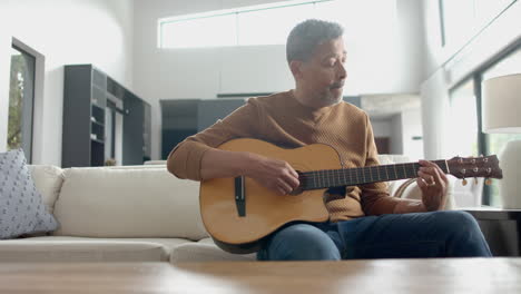 Focused-senior-biracial-man-sitting-on-couch-playing-guitar-at-home,-copy-space,-slow-motion