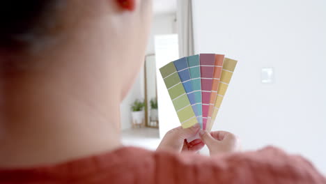 Thoughtful-plus-size-biracial-woman-looking-at-paint-colour-charts-in-sunny-living-room,-slow-motion