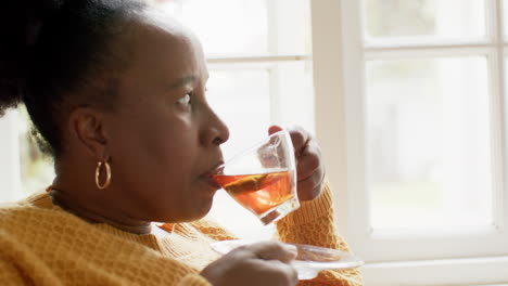 Glückliche-Afroamerikanische-Seniorin,-Die-Tee-Trinkt-Und-In-Einem-Sonnigen-Zimmer-In-Zeitlupe-Nach-Vorne-Blickt