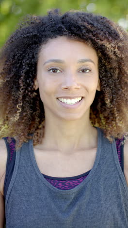 Vertical-video-portrait-of-biracial-woman-with-long-curly-hair-smiling-in-sunny-garden,-slow-motion