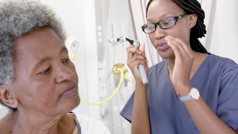 African-american-female-doctor-testing-ear-of-senior-female-patient-in-hospital-room,-slow-motion