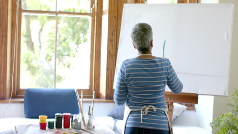 Senior-biracial-woman-wearing-apron-and-painting-on-big-canvas-at-home,-slow-motion