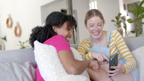 Happy-diverse-teenage-female-friends-talking-and-using-smartphone-on-couch-at-home,-slow-motion