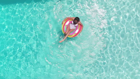 Aerial-of-happy-african-american-teenage-girl-playing-on-inflatable-in-sunny-pool,-copy-space