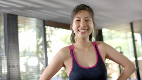 Happy-asian-woman-with-arms-akimbo-looking-at-camera-and-smiling-in-sunny-home-gym,-slow-motion