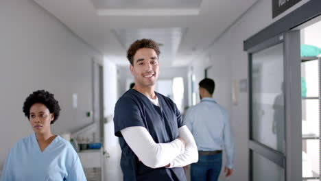 Portrait-of-happy-biracial-male-doctor-wearing-scrubs-in-hospital-corridor,-slow-motion