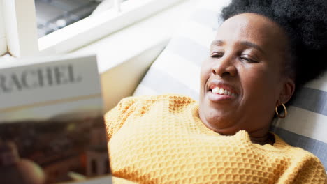 Glückliche-Afroamerikanische-Seniorin,-Die-Im-Sonnigen-Zimmer-Liegt,-Ein-Buch-Liest-Und-Lächelt,-Zeitlupe