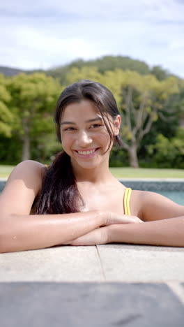 Vertical-video-portrait-of-smiling-biracial-teenage-girl-standing-in-sunny-pool,-slow-motion