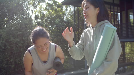Happy-asian-female-friends-with-exercise-mats-and-water-bottle-laughing-on-terrace,-slow-motion