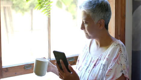 Nachdenkliche-ältere-Frau-Mit-Gemischter-Abstammung,-Die-Tee-Trinkt-Und-Zu-Hause-Am-Fenster-Ihr-Smartphone-Benutzt,-Zeitlupe