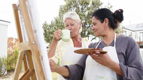 Two-happy-diverse-senior-women-painting-picture-and-smiling-in-sunny-garden,-slow-motion