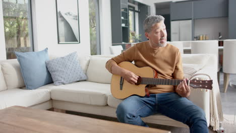 Älterer-Mann-Mit-Gemischter-Abstammung,-Der-Zu-Hause-Auf-Der-Couch-Sitzt-Und-Gitarre-Spielt,-Kopierraum,-Zeitlupe