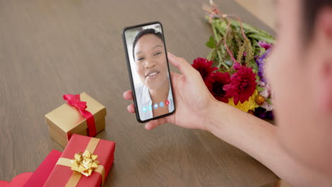 Caucasian-woman-holding-holding-smartphone-with-biracial-woman-on-screen,-gifts-on-desk